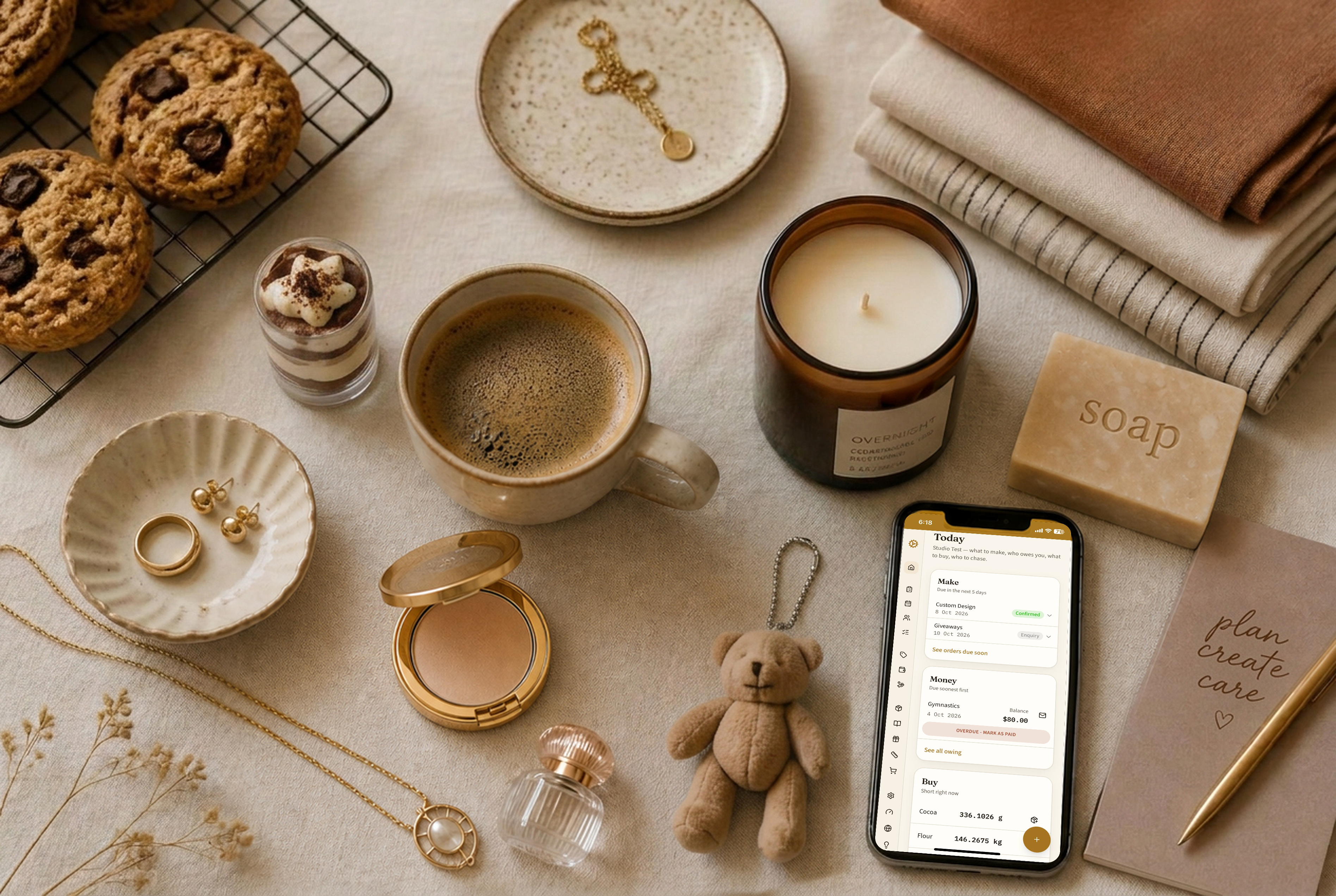 A maker's bench with baked goods, jewellery pliers, folded fabric, a poured candle, and a phone showing an order list